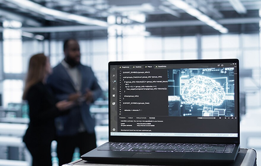 Laptop in a data center displaying code and a glowing digital brain icon with two blurred figures in the background.