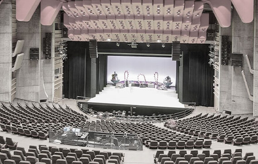Spacious modern concert hall with red ceiling panels, tiered seating, wooden acoustic design, and a brightly lit stage prepared for performance.