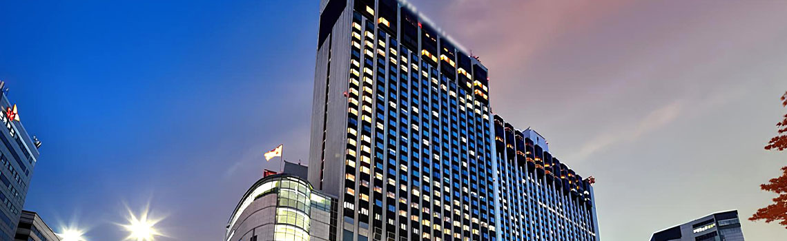 Exterior view of a modern five-star hotel in Myeongdong, Seoul, with illuminated windows and skyline at dusk.
